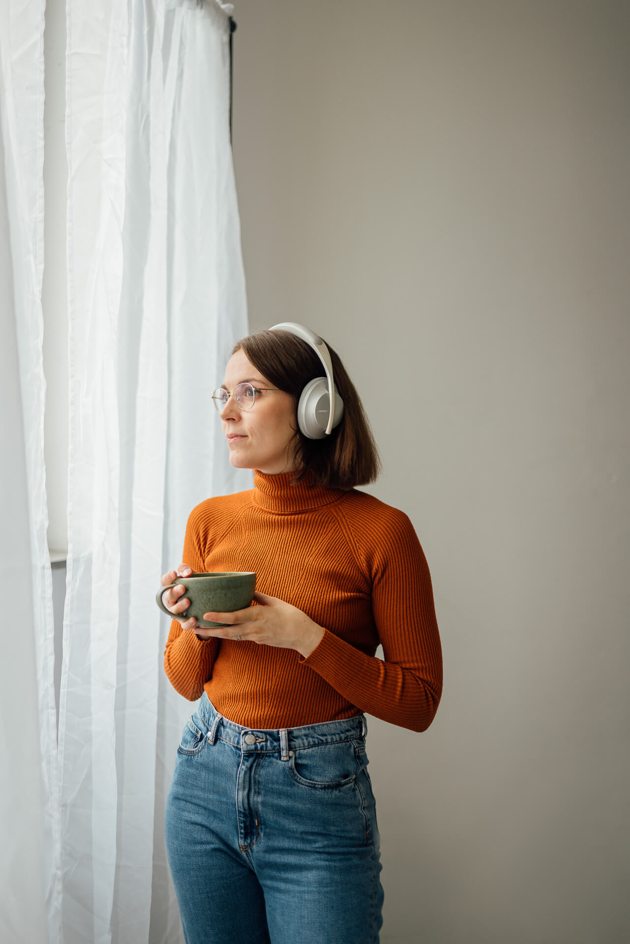 Portrait einer Frau die mit Kopfhörern und einer Tasse Tee am Fenster steht. Sie hört Musik. Das Bild ist während eines Brand Shoots entstanden. 