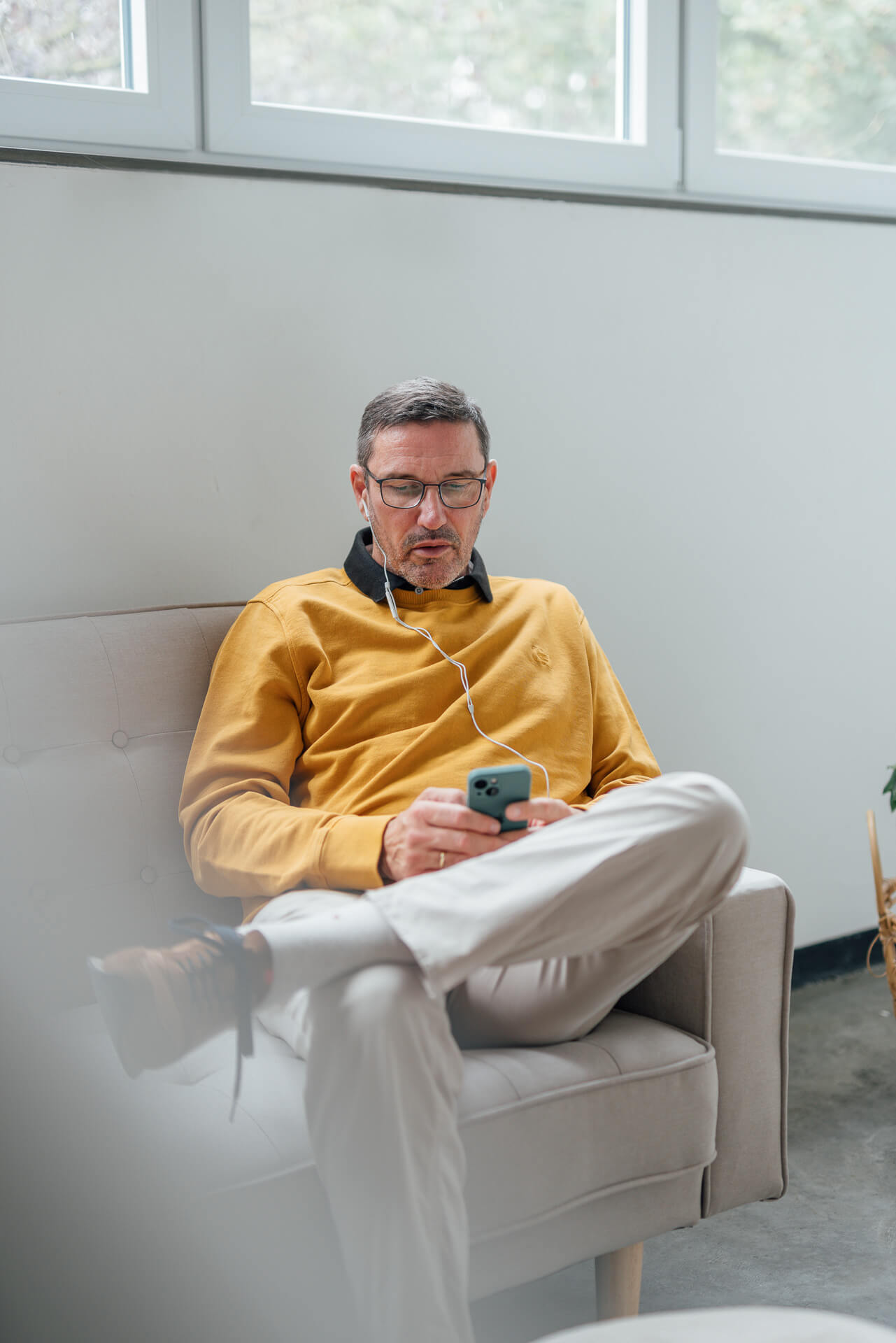 Wolfgang sitzt auf dem Sofa und hat sein Smartphone in der Hand. Er telefoniert und hat Kopfhörer im Ohr. Das Foto ist für ein Brand Shoot gemacht worden