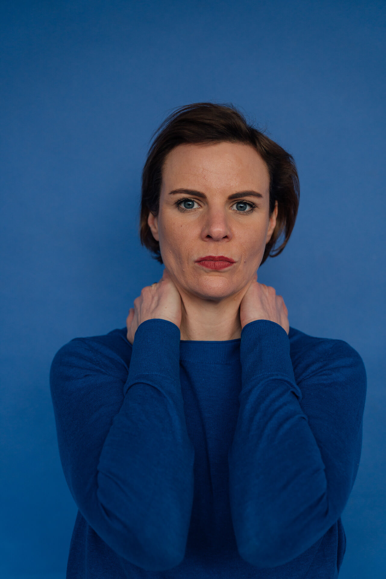 Portrait einer Frau die ihre Hände im Nacken zusammenhält und direkt in die Kamera schaut. Der Hintergrund ist blau und sie trägt einen blauen Pullover. Das Bild ist zum Aufbau einer Personal Brand. 