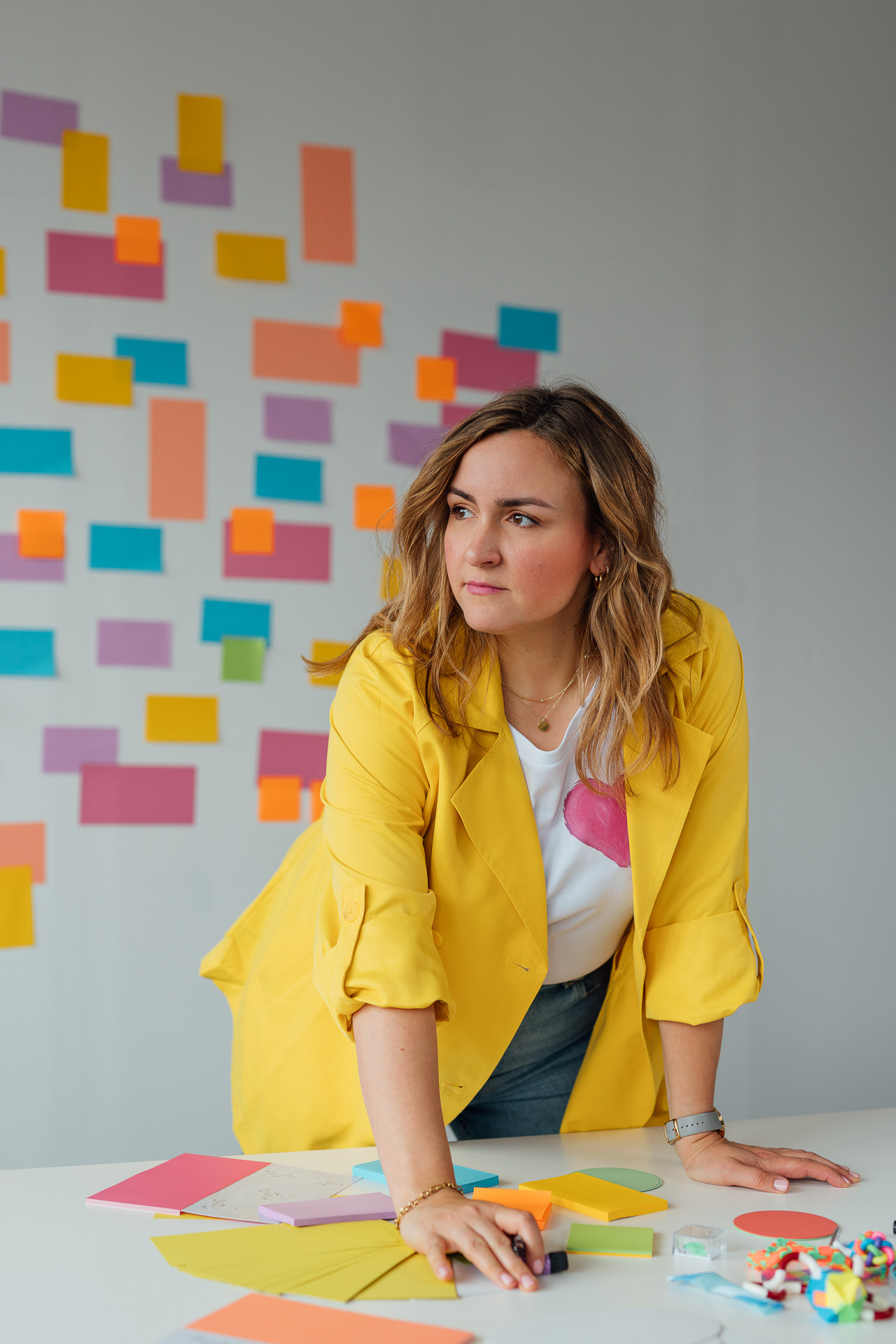 Britt lehnt am Schreibtisch und schaut zur Seite. Das Bild ist ganz bunt, sie hat einen knallgelben Blazer an und im Hintergrund sind ganz viele bunte Postits zu sehen. Das Bild ist für den Aufbau einer Personal Brand auf LinkedIn. 