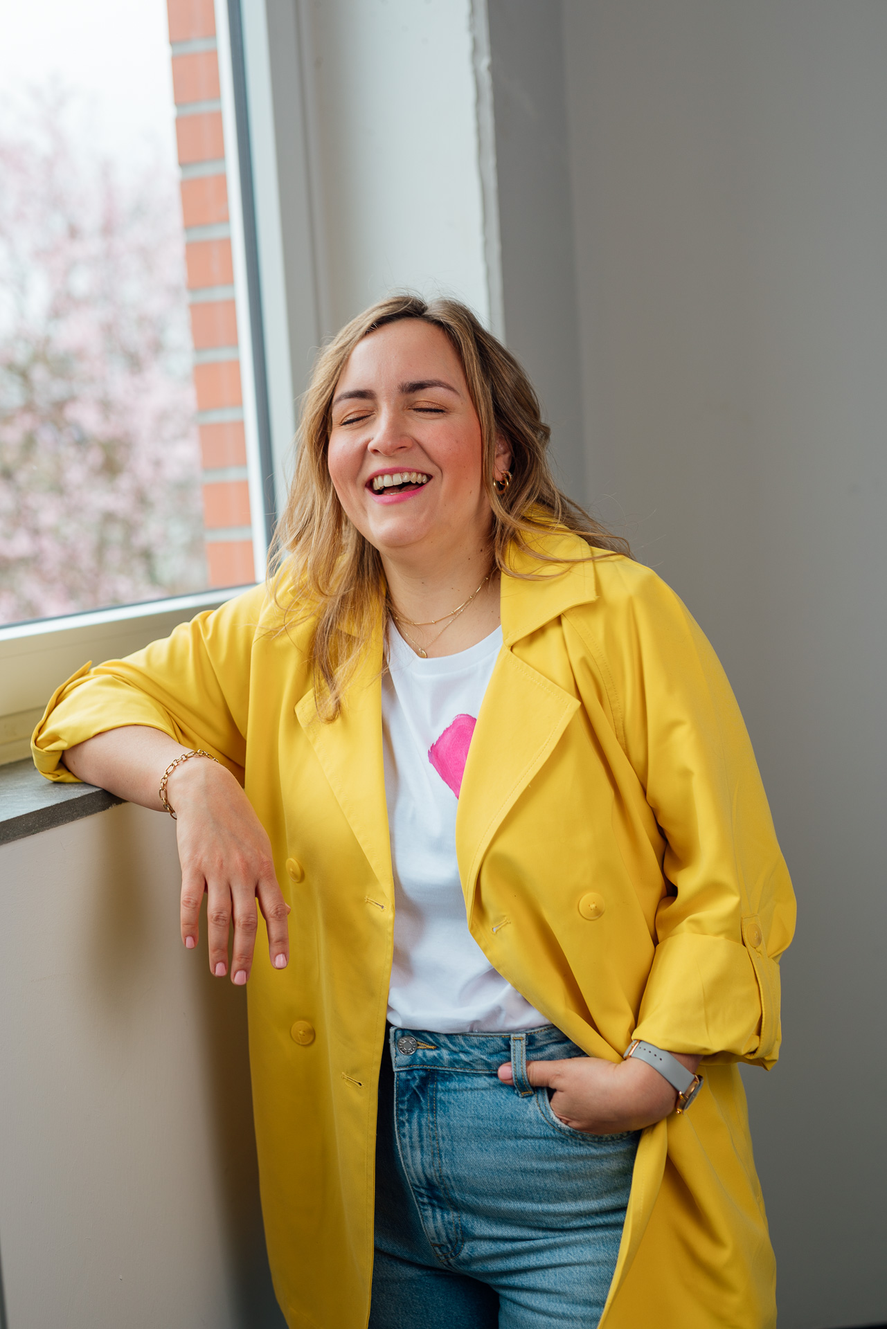 Eine Frau steht mit einem knallgelben Blazer lachend am Fenster und hat dabei einen Arm auf der Fensterbank ablegt. Ein lebenslustiges Foto für eine Personal Brand. 