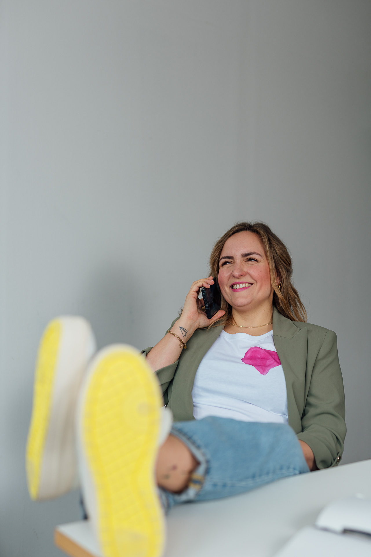 Eine Frau hat die Füße auf dem Schreibtisch abgelegt und telefoniert. Ihre Schuhsolen sind dabei knallgelb. Das Bild ist während eines Brand Shoots entstanden. 