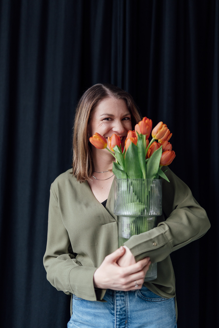 Christin hat eine Vase mit Tulpen in der Hand und steckt ihre Nase rein. Das Bild ist während eines Brand Shoots entstanden und stellt eine natürliche Situation dar. 