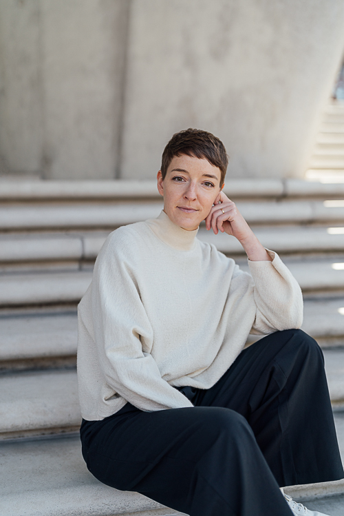 Personal-Branding-Fotos-Hafencity-draußen-Hamburg-1-mobil Businessfoto einer Frau und sie steht in einem beigen Pullover und einer schwarze Hose sitzt in der Hafencity auf einer Treppe.