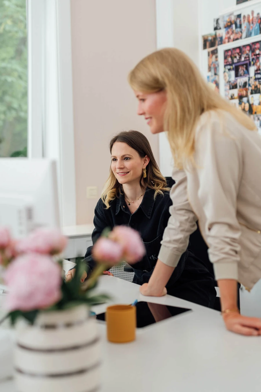 Professionelle Unternehmensfotos aus einem Corporate Shooting in Hamburg: Zwei Frauen arbeiten gemeinsam an einem Computer in einem hellen, modernen Büro, umgeben von persönlichen Details wie Blumen und Bildern, die eine authentische und einladende Arbeitsatmosphäre vermitteln.