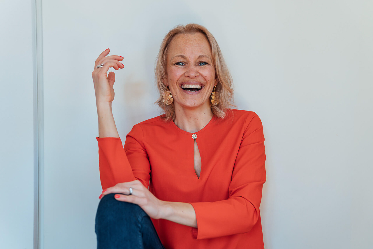 Beispielfoto-Brandingfarben-Brandshoot-2-mobil Branding Portrait einer Frau in einer roten Bluse, sie sitzt auf dem Boden und lacht herzlich in die Kamera