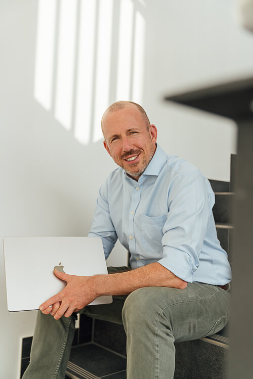 Businessfotos-Geschaeftsfuehrer-Mann-Hamburg-2 Christopher sitzt auf der Treppe mit einem Macbook in der Hand und schaut in die Kamera. Das Bild ist ein sympathisches Businessportrait