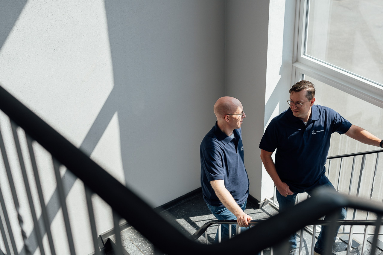 Businessportrait-Baupruefrecht-Hamburg Zwei Männer stehen auf einer Treppe und unterhalten sich. Hinter ihnen an der wand sieht man einen Schatten vom Fenster durch die Sonne. Das Bild ist ein tolles Beispiel für authentische Businessfotos