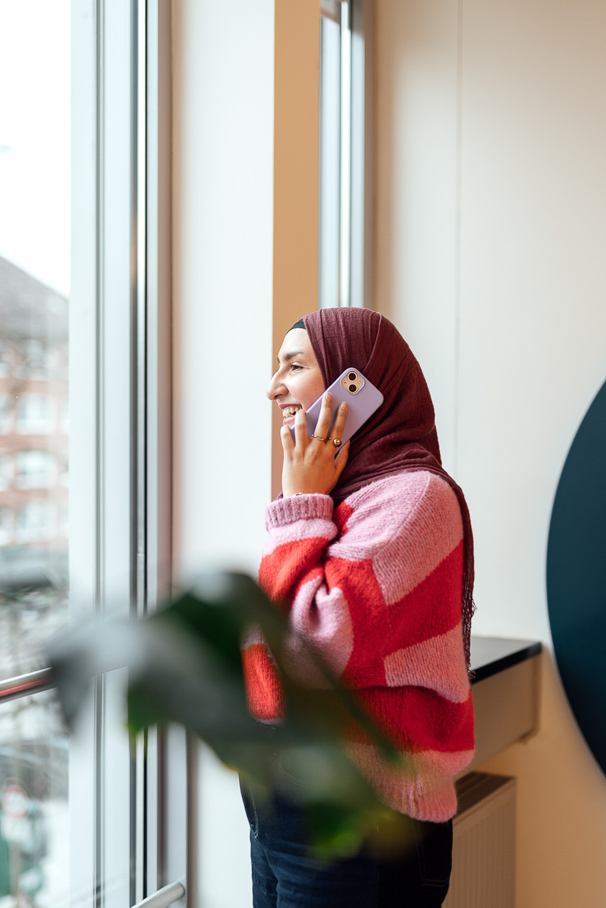 Moodbild-Corporate-Influencer-Content-Bilder-5-mobil Sympathisches Portrait einer Frau, die gerade telefoniert und vor einem Fenster steht, das Bild ist während eines Corporate Influencer Days entstanden.
