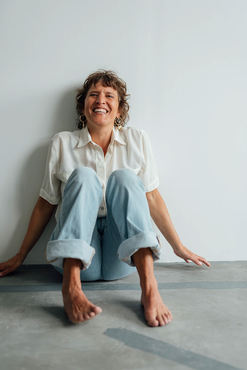 Eine Frau mit weißer Bluse und blauer Jeans sitzt für ein Branding Foto auf dem Boden und lacht in die Kamera