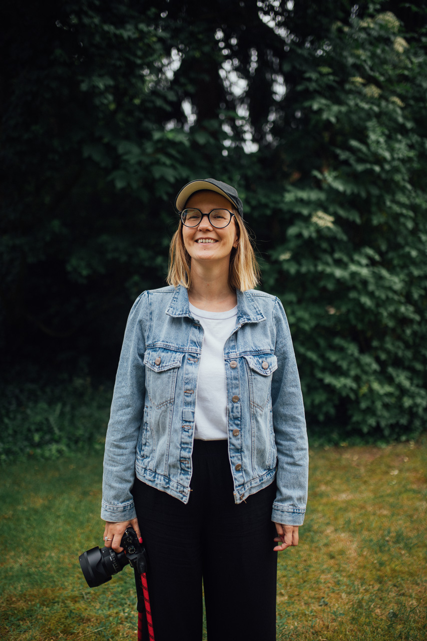Mirjam Kilter-Brandingfotografin-ueber-mich-Seite-mobil Personal Branding Fotografin Mirjam Kilter steht auf einer Wiese. Hinter hier sind grüne Büsche, sie hat eine Kamera in der Hand und ein Cappi auf.