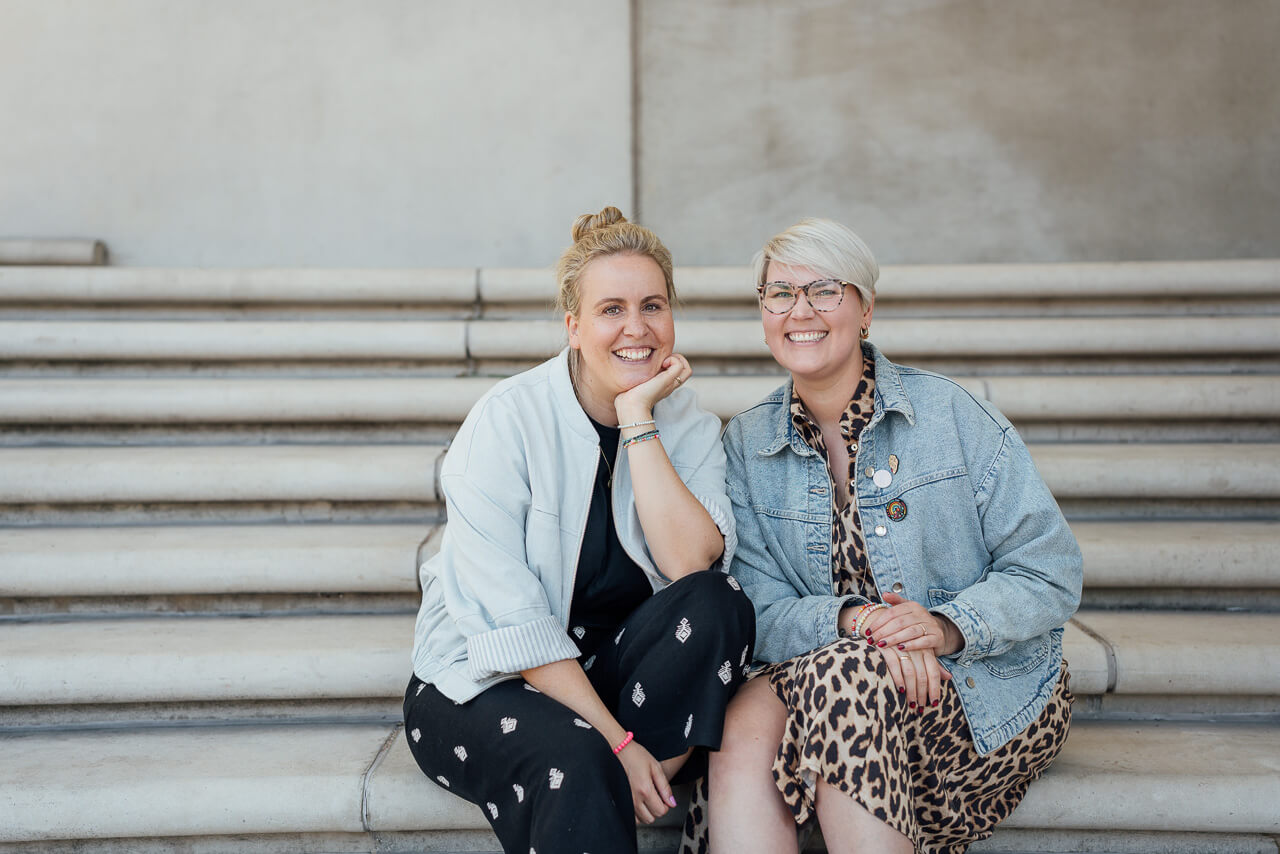 Melanie und Sonja sitzen auf der Treppe und schauen in die Kamera. Das Bild ist während eines Brand Shoots entstanden.  