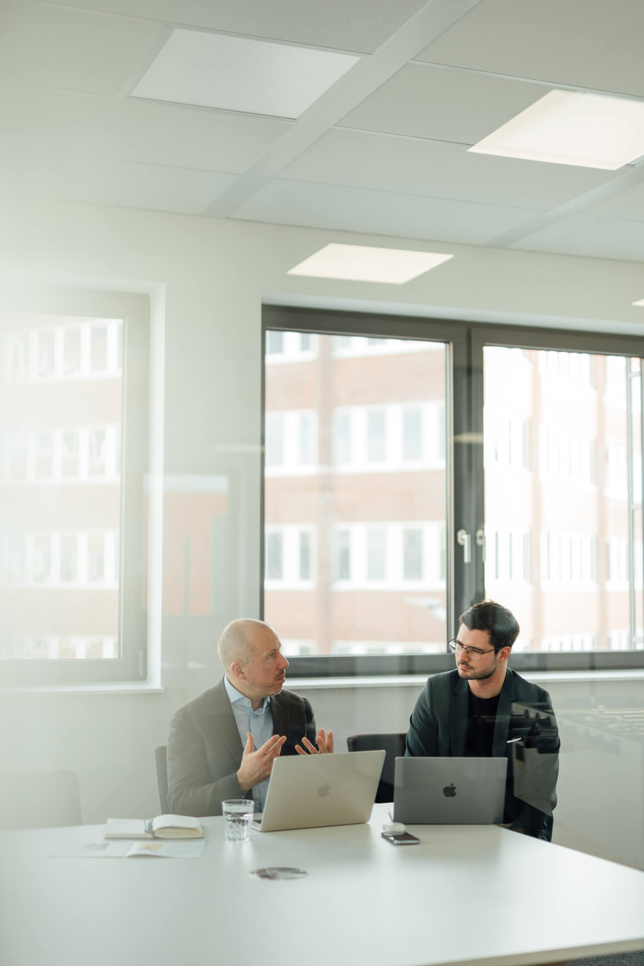 Businessfoto-mittelstandisches-Unternehmen-Hamburg-1 Meetingsituation mit Christopher, der sich mit einem Kollegen unterhält. Beide sitzen am Tisch und es steht ein Laptop auf dem Tisch.