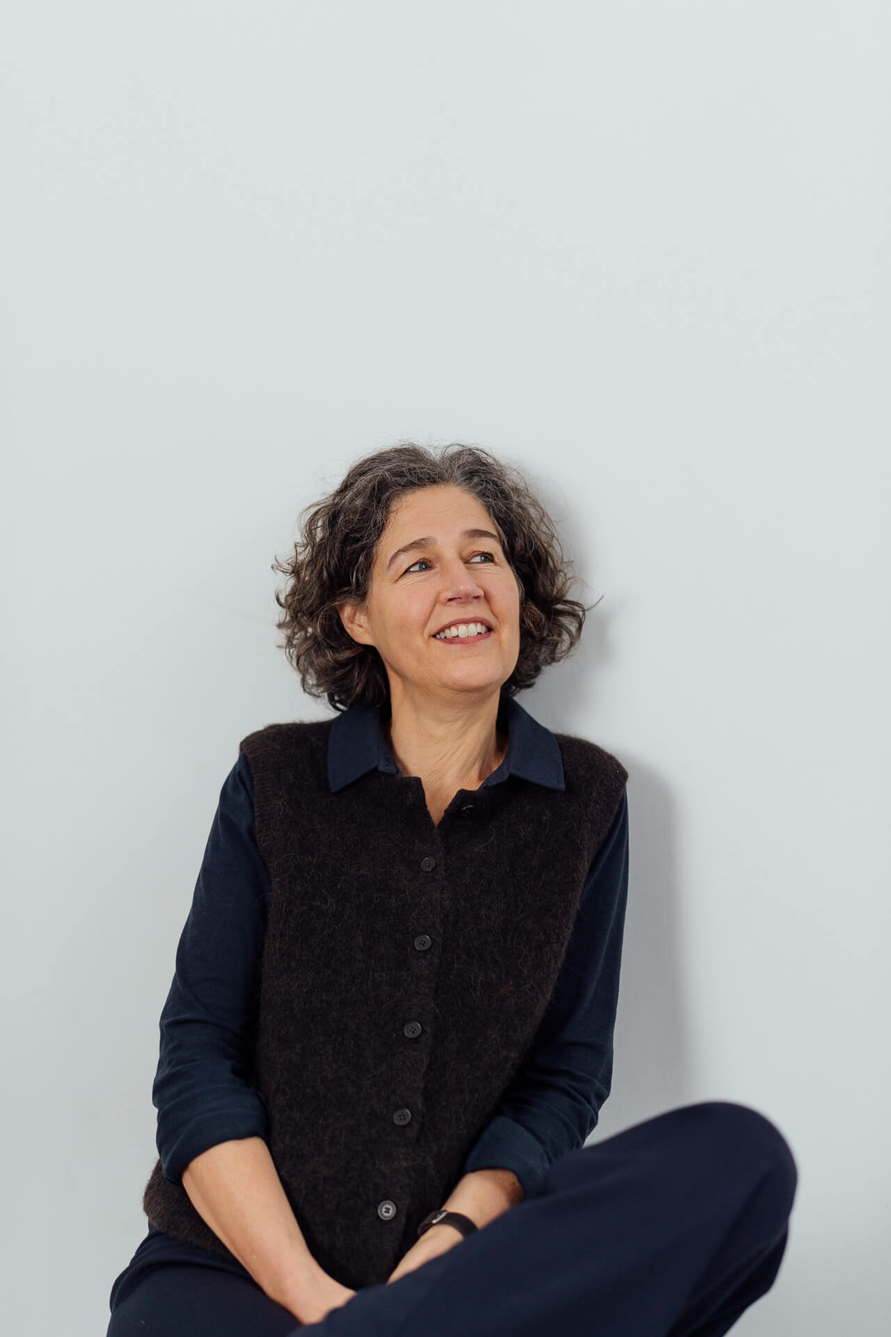 Portrait einer Frau, die in einem Hosenanzug aus Jeans vor einem grünen Hintergrund steht und leicht zur Seite lächelt