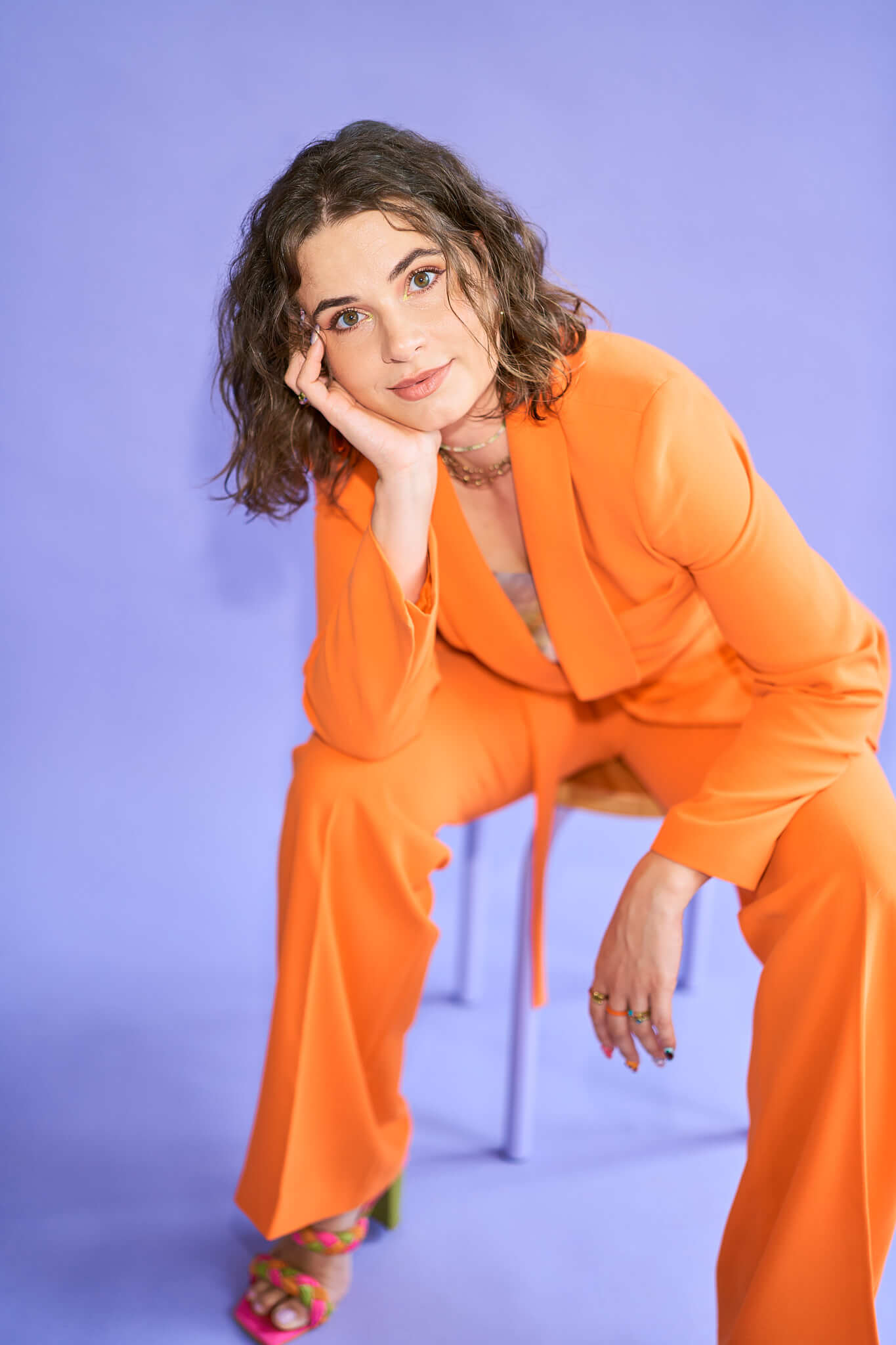 Personal-Branding-Fotografie-Hamburg-Portrait-Frau-1 Antonia sitzt auf einem Hocker leicht nach vorne gelehnt auf ihre Rechte Hand, sie hat ein knallorangenes Outfit an und sitzt vor einem lila Hintergrund
