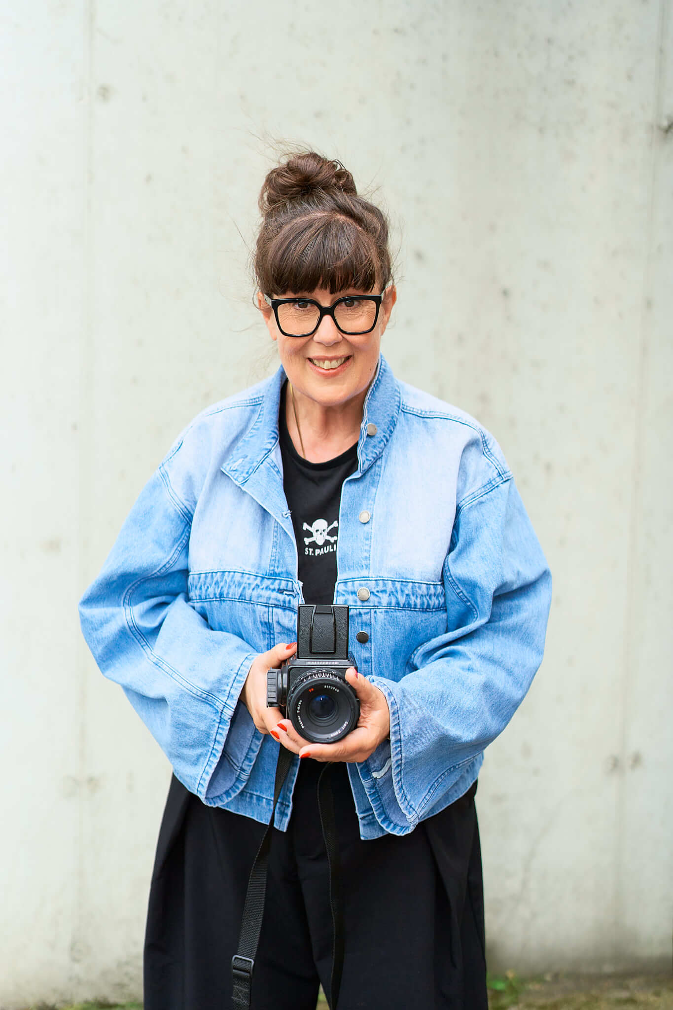Personal-Branding-Fotografie-Hamburg-Portrait-einer-Fotografin Portrait von Kerstin, die mit einer Jeansjacke und schwarzer Hose direkt in die Kamera schaut, sie hat eine Hasselblad in der Hand und steht vor einer beigen Mauer draußen