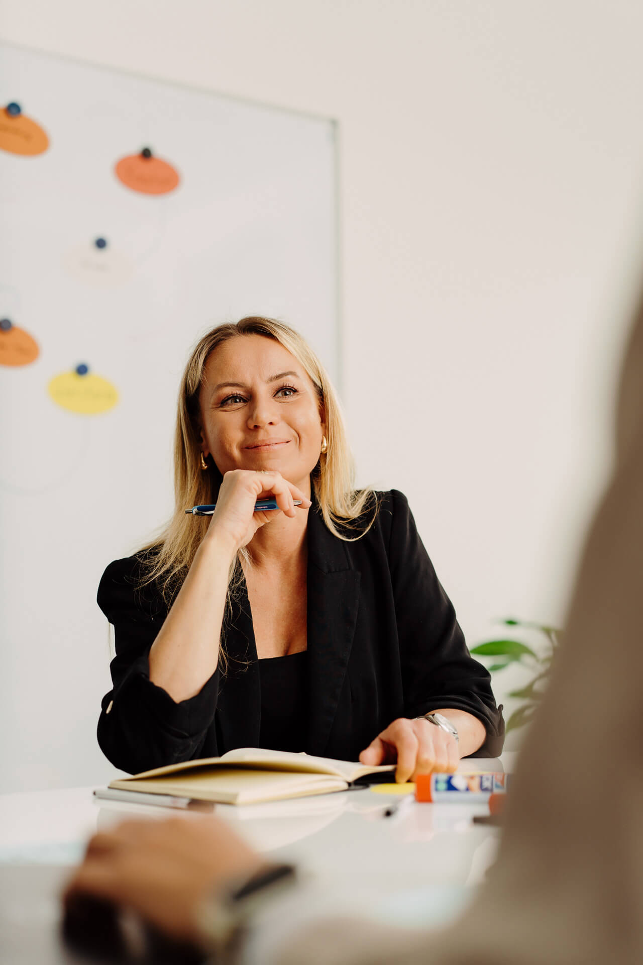 Portrait-Mitarbeiterin-Corporate-Business-Shooting-1 Eine Frau sitzt an einem Tisch und hat den rechten Arm an ihrem Kinn, hinter ihr sind Moderationskarten an die Wand geklebt worden und vor ihr sitzt ein Kollege an dessen Schulter vorbeifotografiert wurde