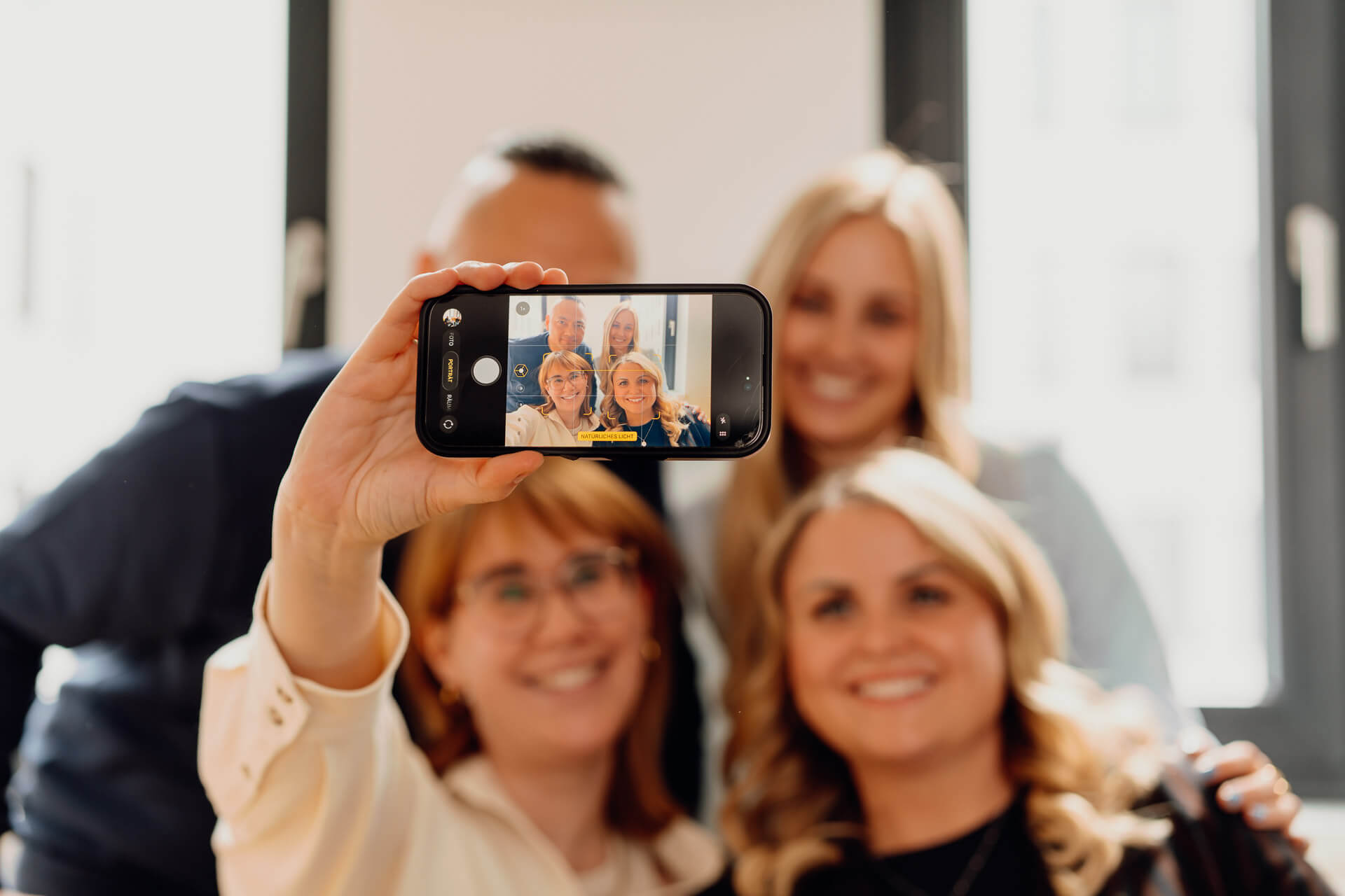 Selfie-Business-Shooting-Businessfotografie-Hamburg Eine Frau hält das Smartphone in ihrer Hand und macht ein Selfie mit ihren Kollegen, im Rahmen eines Business Shootings, genau dieses Motiv wird gerade fotografiert