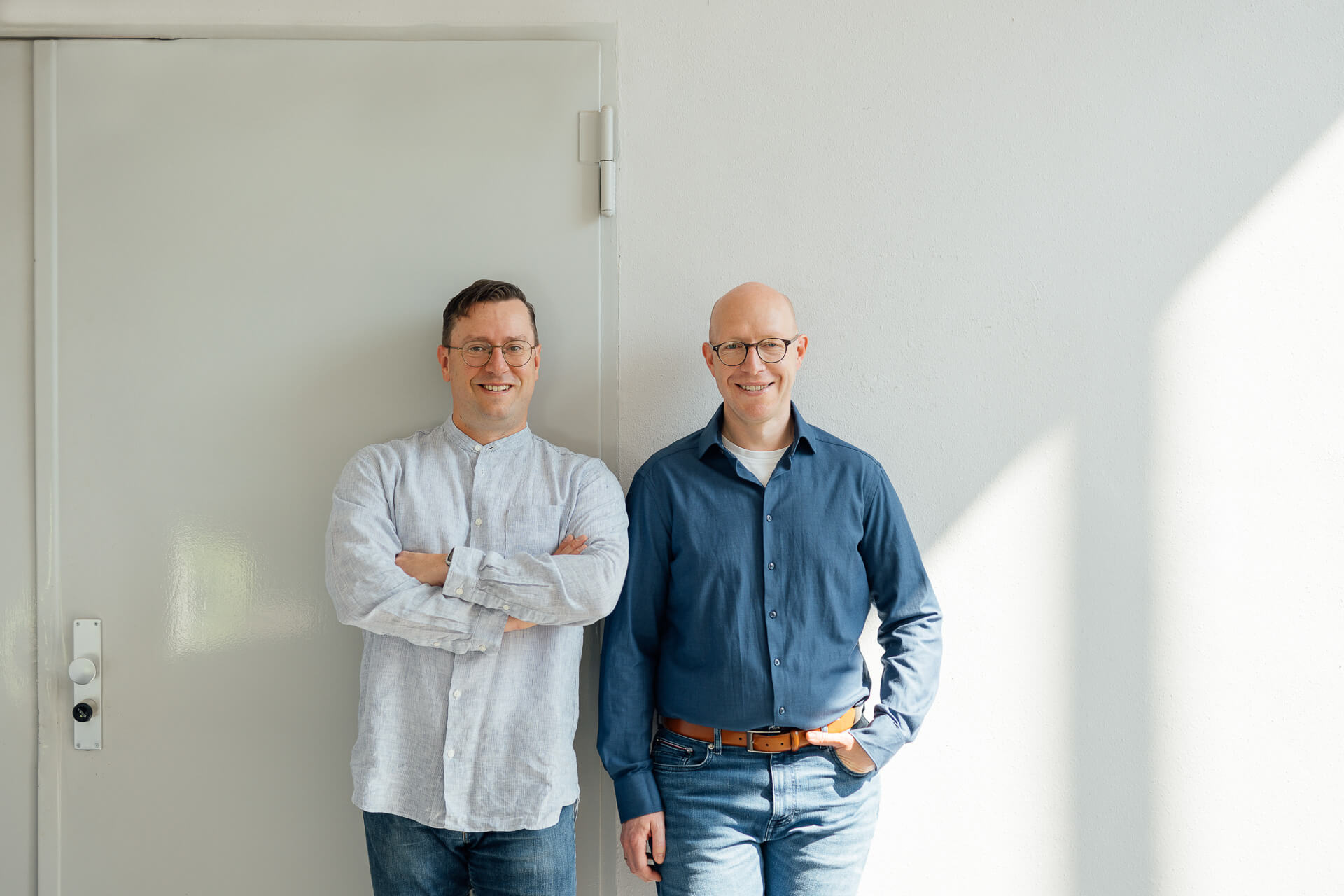 Businessfoto-Portraits-Geschaeftsfuehrer-Hanse-Inspect Teamportrait von zwei Geschäftsführern der Hanse Inspect GmbH, die lächelnd nebeneinander an einer hellen Wand im Sonnenlicht stehen