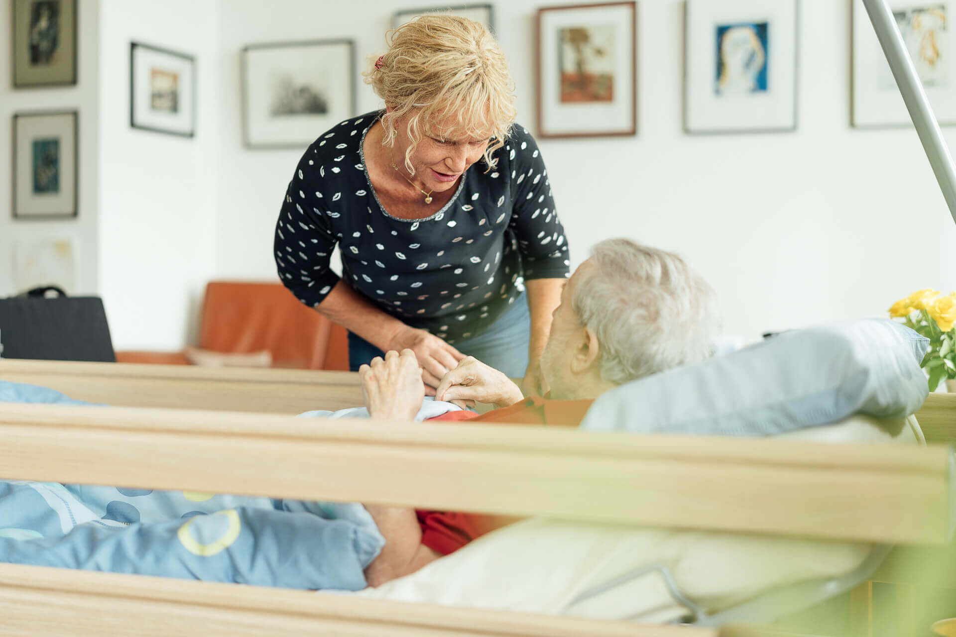 businessfoto-haeusliche-betreuung-bett Pflegekraft in der häuslichen Betreuung beugt sich zu einem älteren Mann im Pflegebett hinunter
